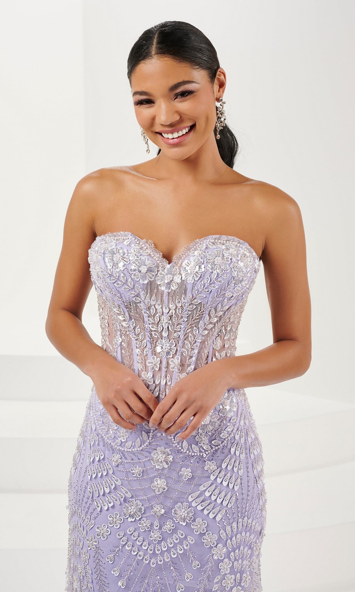 A woman stands and smiles in the Tiffany Strapless Beaded Mermaid Prom Dress 16103, a lavender gown with floral beading, against a plain white background.